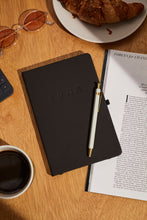 Load image into Gallery viewer, LDNA London black notebook on a wooden table alongside a pen, croissant, glasses, coffee and magazine.