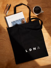 Load image into Gallery viewer, LDNA London black tote bag on a wooden table with lifestyle items such as magazine, coffee, sunglasses and pen.